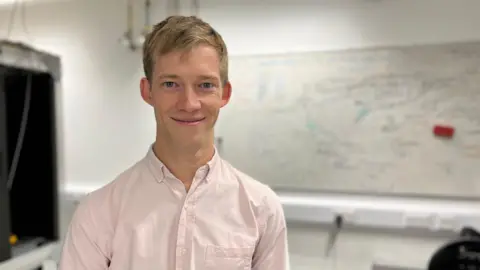 BBC A blonde man in a pale pink shirt standing in front of a whiteboard with writing on it