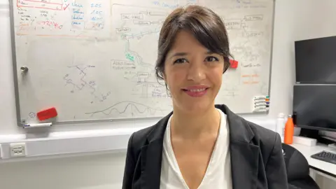 A woman with dark hair and wearing a white shirt and a black blazer smiling at the camera with a whiteboard behind her